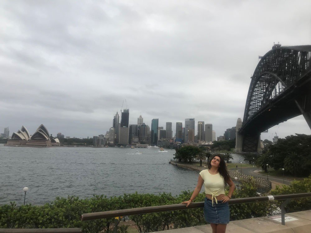 Experiencia estudiante chica morena con una camiseta amarilla y una falta tejana, posando delante de la ciudad de Sydney