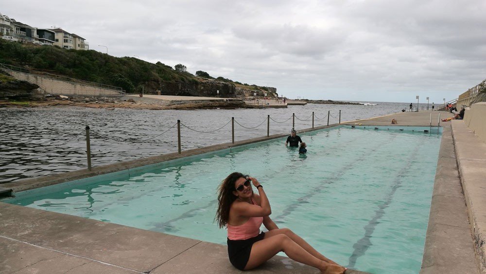 Vivir en Sydney chica morena posando sentada en las piscinas de Bondi Beach en Sydney. De fondo el cielo nublado