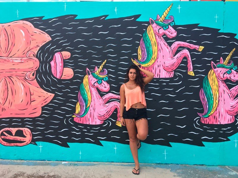 Vivir en Sydney chica morena posando en una pared pintada con unicornios rosas. Ella lleva una camiseta de tirantes naranja y un pantalón corto negro