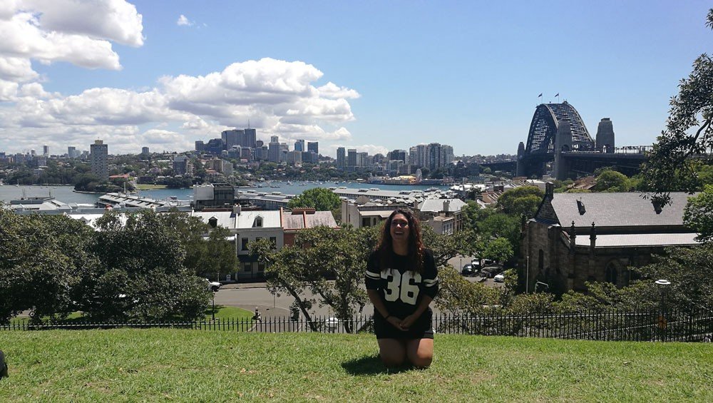 Vivir en Australia chica morena de rodillas en el césped y de fondo la ciudad de Sydney con el cielo despejado